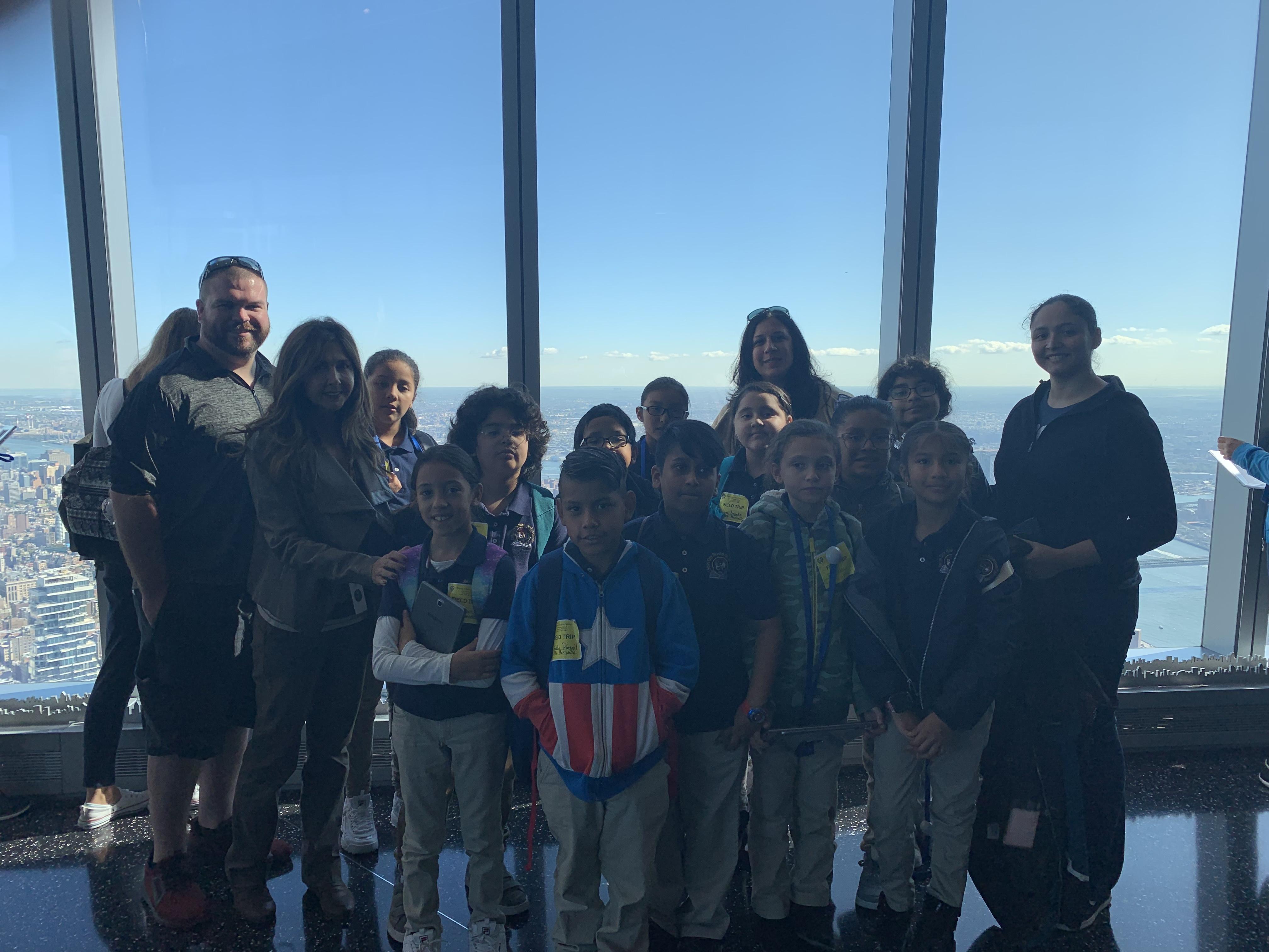Mr. Blake, Marinello, Ms. Logothetis, & Ms. Cintron and several students standing in front of the window with the NYC background