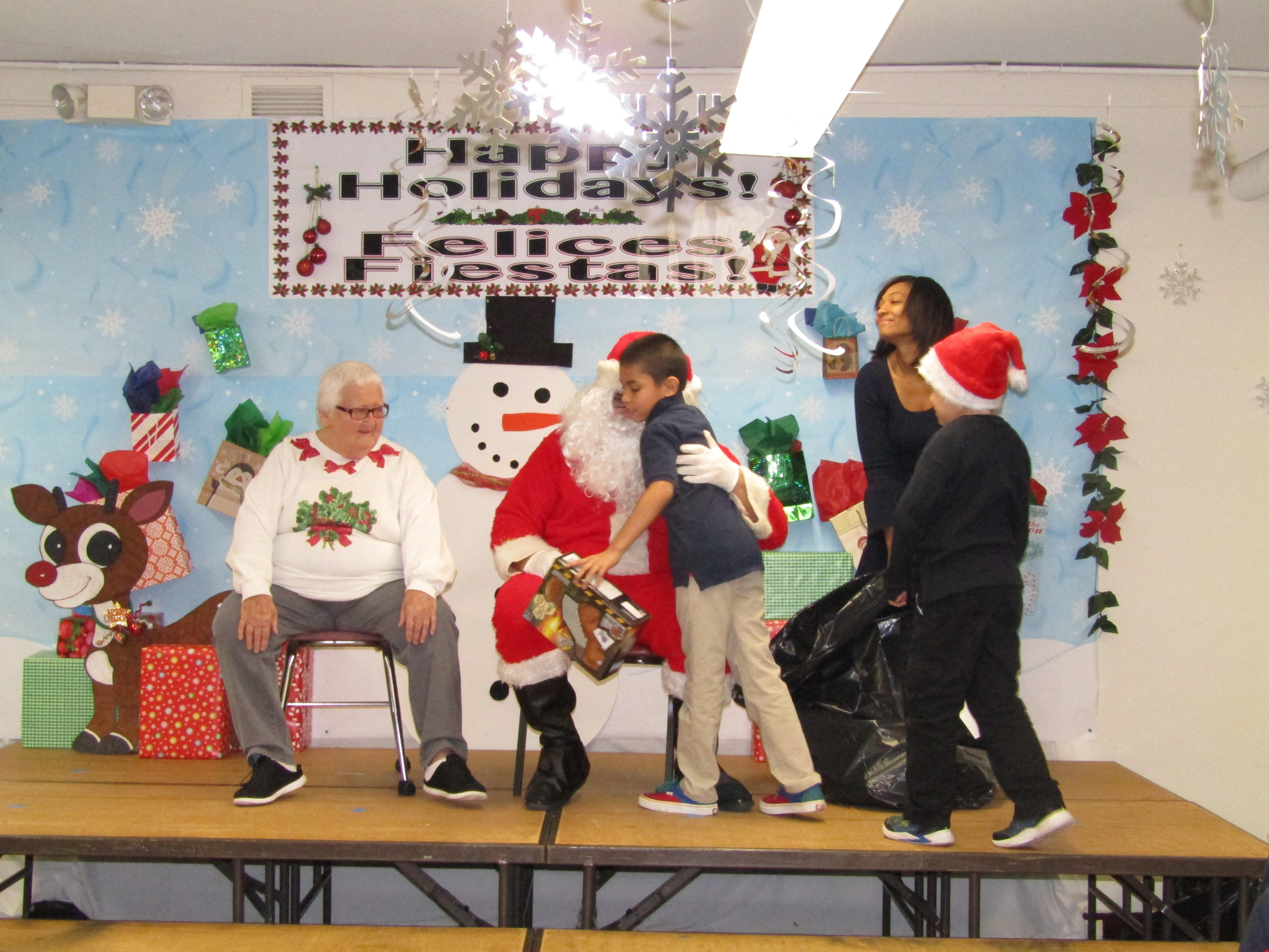 Mr. & Mrs. Claus handing out gifts to male student on stage as teacher pulls gift from bag