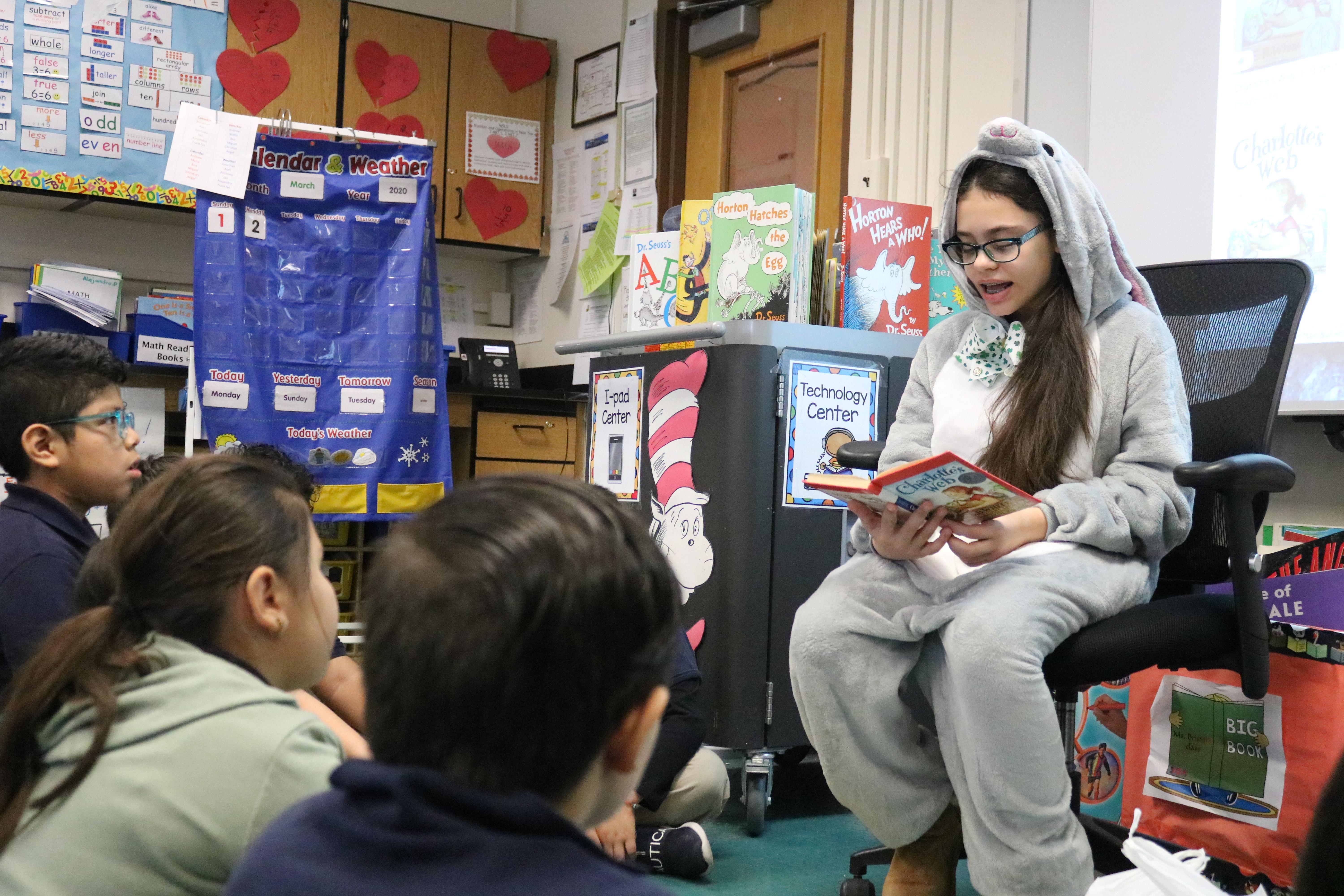 Girl with glasses wearing hooded pj reading a book to Ms. Bristol's Class close up