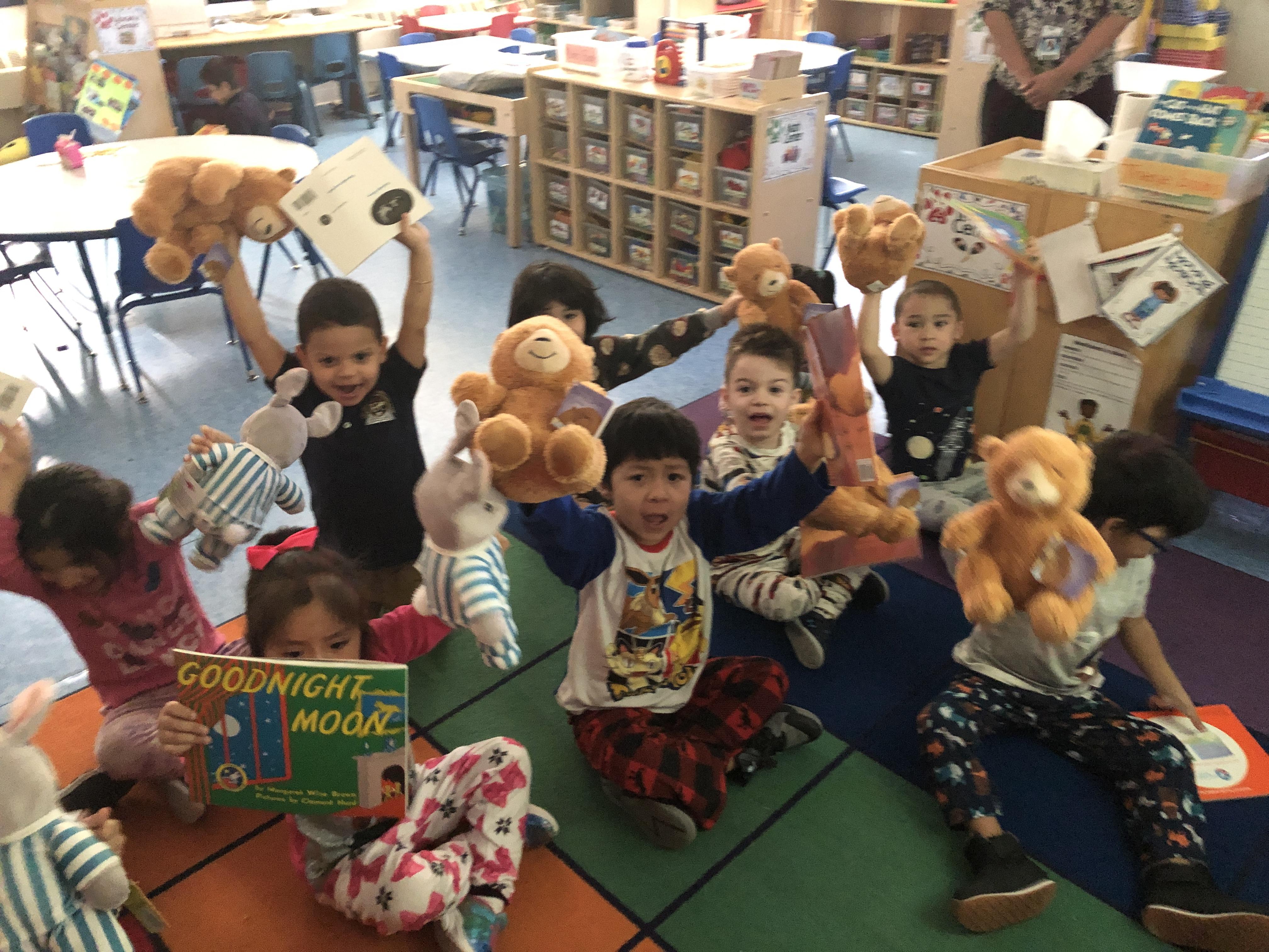 kids wearing pj's sitting on the class rug happily holding up their books and stuffed animals friends